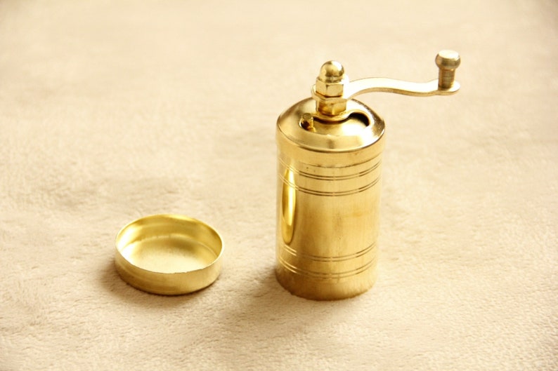 May include: A gold metal pepper grinder with a crank handle. The grinder has a cylindrical shape and is decorated with horizontal lines.
