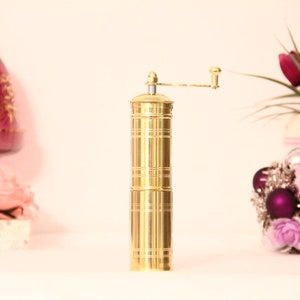 May include: A gold-colored pepper grinder with a hand crank. The cylindrical grinder has horizontal etched lines and a small handle. Decorative elements include a pink glass goblet and a floral arrangement with purple and silver ornaments.