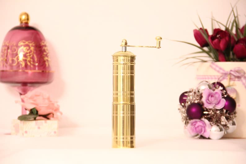May include: A gold-colored pepper grinder with a hand crank. The cylindrical grinder has horizontal etched lines and a small handle. Decorative elements include a pink glass goblet and a floral arrangement with purple and silver ornaments.
