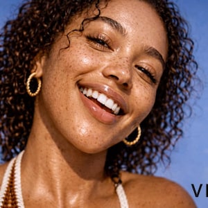 Puede incluir: Una persona sonriente con cabello casta&ntilde;o rizado y pecas lleva pendientes de aro dorados. La persona lleva un top halter blanco y marr&oacute;n. La palabra "VIVRE" est&aacute; en texto blanco en la esquina inferior derecha.
