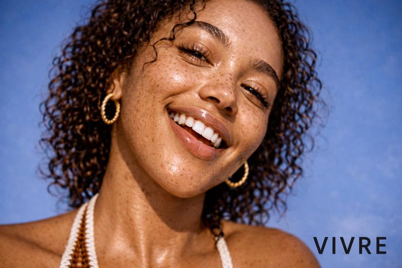Puede incluir: Una persona sonriente con cabello casta&ntilde;o rizado y pecas lleva pendientes de aro dorados. La persona lleva un top halter blanco y marr&oacute;n. La palabra "VIVRE" est&aacute; en texto blanco en la esquina inferior derecha.