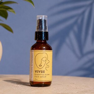 Puede incluir: Una botella de vidrio marr&oacute;n de s&eacute;rum facial con una bomba negra y una etiqueta amarilla. La etiqueta presenta la palabra "VIVRE" y el texto "ESSENTIAL GLOW FACE SERUM". La botella est&aacute; sobre una superficie clara con un fondo azul.
