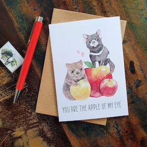May include: A greeting card featuring a watercolour illustration of a Tasmanian devil and a wombat holding apples. The card reads "You are the apple of my eye."
