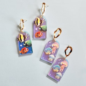 Könnte beinhalten: Ein Paar Ohrhänger mit einer goldfarbenen Metalleinfassung. Die Ohrringe haben einen regenbogenfarbenen Hintergrund in Lila und Blau mit einem Design aus bunten Fischen und Quallen.