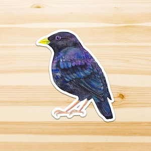 Op de afbeelding: Een gestanste sticker met een gedetailleerde aquarelillustratie van een spreeuw. De vogel heeft iriserende zwarte, blauwe en paarse veren, een heldergele snavel en lichtbruine poten. De sticker is voorzien van een dunne witte rand.