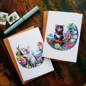 May include: Two greeting cards with watercolor illustrations. One card features a koala sitting on a branch surrounded by flowers and leaves. The other card features a Tasmanian devil standing on a branch with flowers and leaves.
