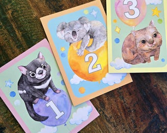 May include: Three rectangular greeting cards featuring watercolor illustrations of Australian animals. One card shows a black Tasmanian devil with the number 1, another a koala with the number 2, and the third a wombat with the number 3 on a balloon.