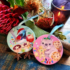 May include: Two circular Christmas decorations with watercolour illustrations. One depicts a bird in a Santa hat, an apple, and pastries on a light blue backdrop. The other shows a wombat with a gingerbread man and cookies on a pink background.