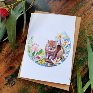 May include: A watercolor illustration of a wombat sitting in a circle of flowers and leaves. The wombat is brown and has a white nose and belly. The flowers are pink, yellow, and white. The leaves are green.