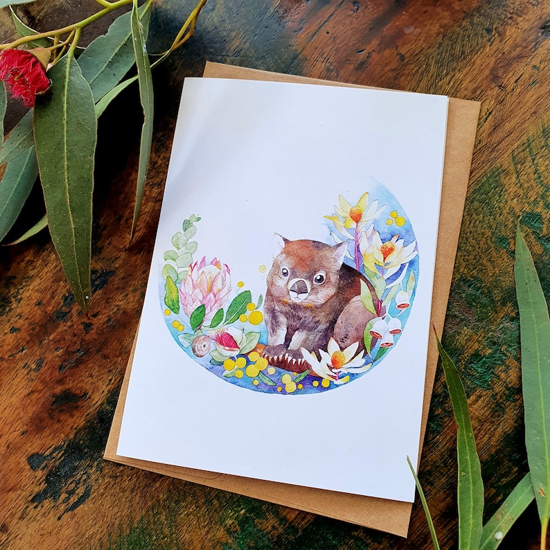 May include: A watercolor illustration of a wombat sitting in a circle of flowers and leaves. The wombat is brown and has a white nose and belly. The flowers are pink, yellow, and white. The leaves are green.