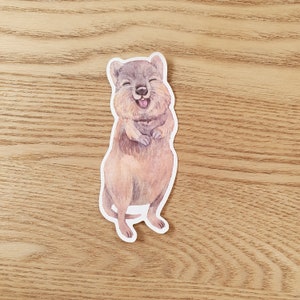 May include: A watercolor illustration of a quokka, a small marsupial native to Rottnest Island, Australia. The quokka is smiling with its mouth open, showing its teeth. The quokka is standing on its hind legs with its front paws raised.