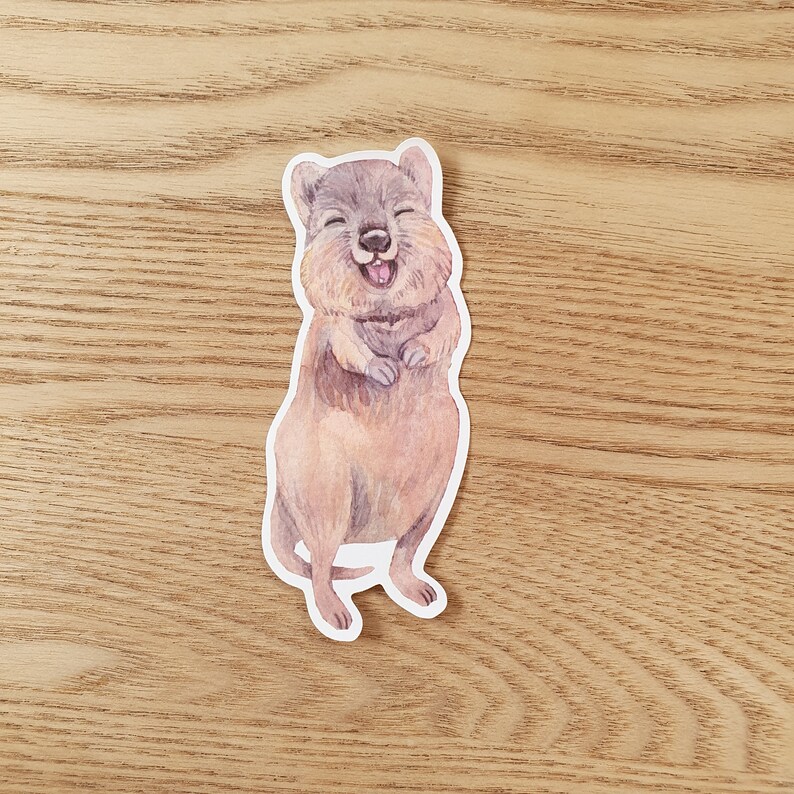 May include: A watercolor illustration of a quokka, a small marsupial native to Rottnest Island, Australia. The quokka is smiling with its mouth open, showing its teeth. The quokka is standing on its hind legs with its front paws raised.
