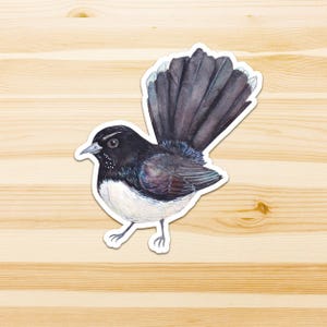 Op de afbeelding: Een gestanste sticker met een aquarelillustratie van een waaierstaartvogel. De vogel heeft een zwarte kop, vleugels en een grote, waaiervormige staart met witgepuntte veren. De vogel heeft een witte borst en buik met dunne, lichtgrijze poten.