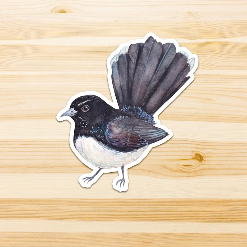 Op de afbeelding: Een gestanste sticker met een aquarelillustratie van een waaierstaartvogel. De vogel heeft een zwarte kop, vleugels en een grote, waaiervormige staart met witgepuntte veren. De vogel heeft een witte borst en buik met dunne, lichtgrijze poten.