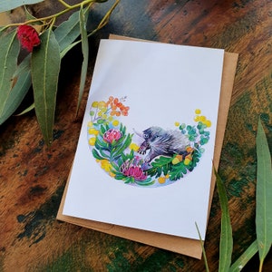 May include: A watercolor illustration of an echidna surrounded by yellow, green, and pink flowers. The echidna is in a curled up position with its back facing the viewer. The illustration is on a white card with a brown envelope.