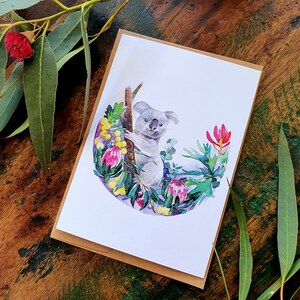 May include: A watercolor illustration of a koala sitting in a tree with pink, yellow, and green flowers. The koala is gray and has a white chest. The background is a light blue.