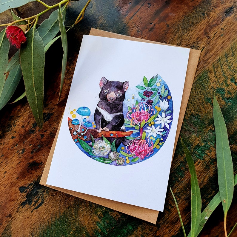 May include: Watercolor illustration of a Tasmanian devil sitting on a branch surrounded by flowers and mushrooms. The devil is black with a white chest and a pink nose. The flowers are pink, purple, and white. The mushrooms are blue and white.