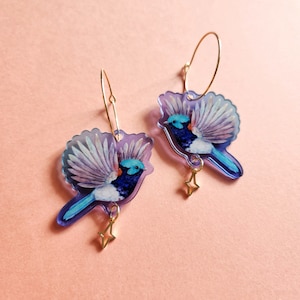 Könnte beinhalten: Ein Paar Ohrringe in Form von blauen und violetten Vögeln mit goldenen Kreisen und Sternanhängern. Die Vögel haben weiße und blaue Flügel und einen blauen Körper mit einem roten Streifen.