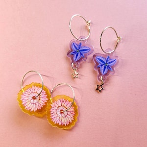 Puede incluir: Dos pares de pendientes de aro dorados con diseños florales. Un par presenta una flor morada con un dije de estrella, y el otro par presenta una flor rosa.