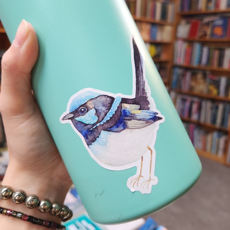 Op de afbeelding: Een gestanste sticker met een aquarelillustratie van een kleine vogel met een felblauwe kop, een donkerblauwe en zwarte borstband en een witte buik. De vogel heeft een lange, rechtopstaande staart, donkere vleugels en dunne, lichtbruine poten.
