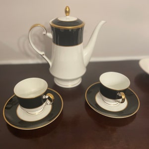 May include: A white coffee pot with a black and gold trim and two matching cups and saucers.
