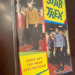May include: A vintage Star Trek comic book, sealed in plastic. The cover features images of the Star Trek crew in their uniforms. The comic book has a yellow banner with the words "STAR TREK" and "GOLD KEY 100-PAGE SPECTACULAR".