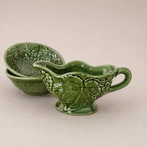 May include: Green ceramic serving set featuring a gravy boat and two small bowls. The pieces have a grape and leaf design. The gravy boat has a handle and a spout. The bowls are stacked. The set is on a neutral background.