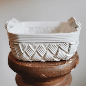 May include: A white ceramic basket with a rectangular shape and a woven texture pattern on the exterior. The basket features a smooth, flat rim and two small, integrated handles on opposite sides, crafted with a glossy, uniform white finish.
