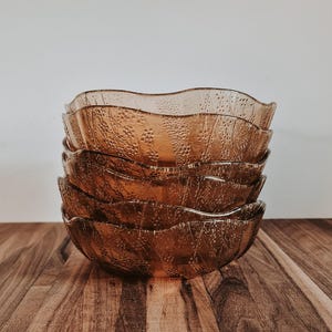 May include: Five amber-coloured glass bowls with a textured pattern, stacked on a wooden surface. The bowls have a wavy rim. The background is a plain white wall. These bowls are suitable for serving food. The bowls are likely to be measured in centimetres.
