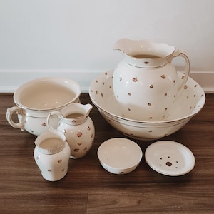 May include: A six-piece ceramic washbasin set in cream with a delicate floral pattern and gold trim. The set includes a large pitcher and basin, a chamber pot, a small pitcher, a small bowl, and a soap dish with a perforated insert for drainage.