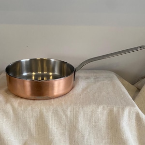 Puede incluir: Una cacerola con fondo de cobre, interior de acero inoxidable y un mango largo plateado. La sartén redonda se encuentra sobre una superficie clara y texturizada. El mango se extiende hacia la derecha.