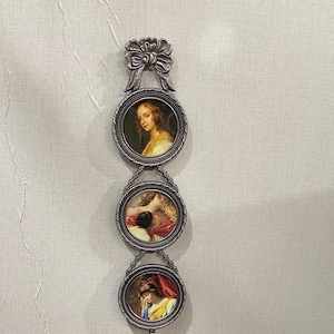 Può includere: Una decorazione da parete con tre ritratti ovali di donne in cornici argentate ornate. Le cornici sono collegate da elementi decorativi e sormontate da un fiocco. Una nappa pende dal basso.