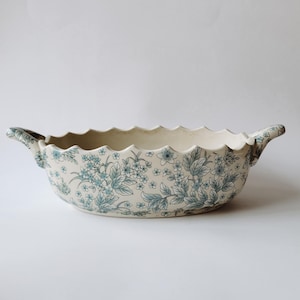 Peut inclure: Un bol de service en céramique couleur crème doté d'un bord festonné et de deux poignées latérales. L'extérieur est orné d'un motif floral bleu répétitif composé de petites fleurs et de branches feuillues couvrant toute la surface.