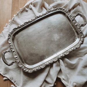 Puede incluir: Una bandeja de servicio rectangular de tono plateado con detalles florales en relieve a lo largo de los bordes elevados y dos asas decorativas en los extremos. La superficie central de la bandeja muestra un patrón grabado sutil.