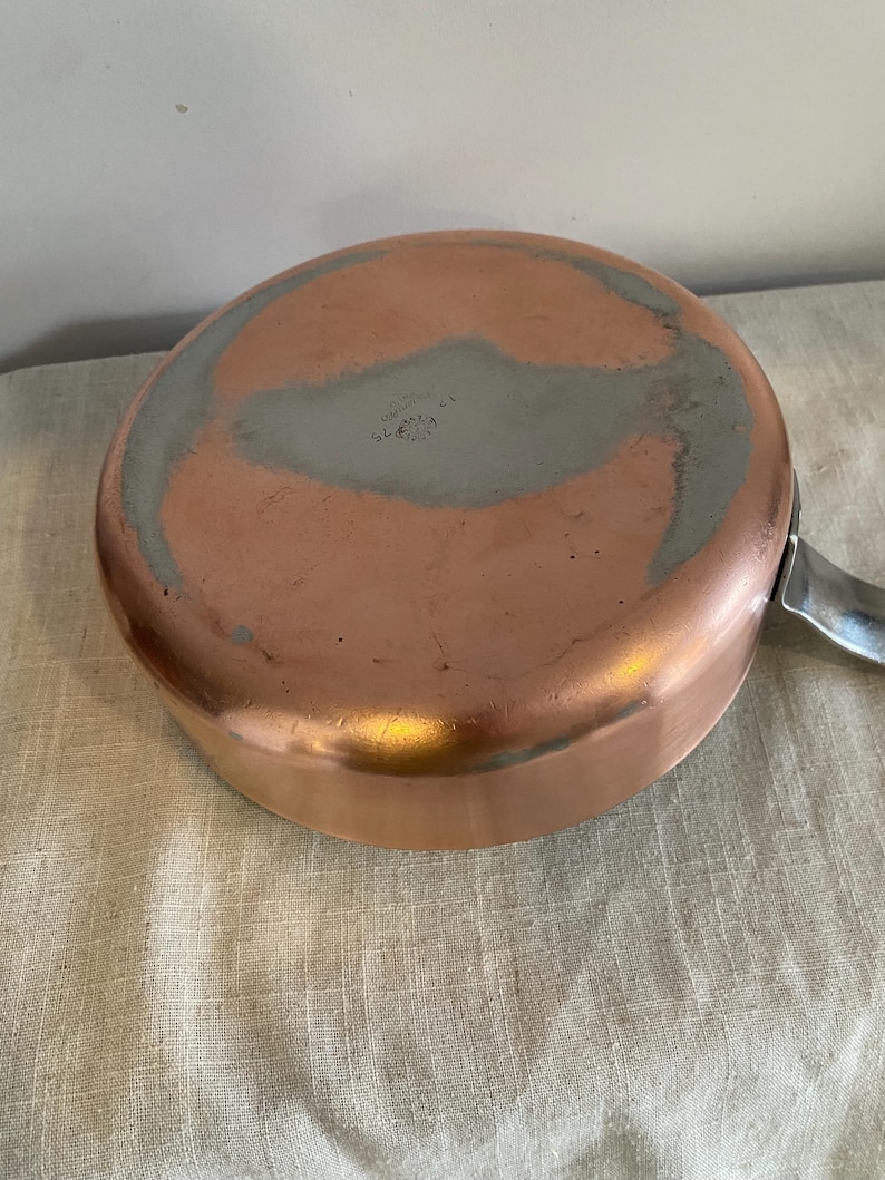 May include: A copper saucepan with a silver handle. The base of the pan shows wear and a grey interior coating. This pan is likely used for cooking and is a kitchenware item.