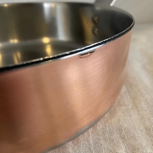 May include: Close-up of a copper saucepan with a stainless steel interior and a silver handle. The pan has a rounded base and a slightly flared rim. The handle is attached with rivets.