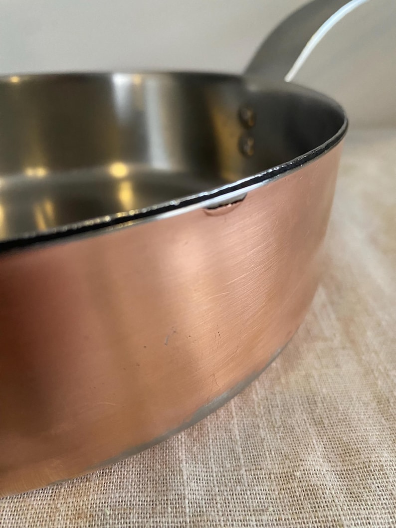 May include: Close-up of a copper saucepan with a stainless steel interior and a silver handle. The pan has a rounded base and a slightly flared rim. The handle is attached with rivets.