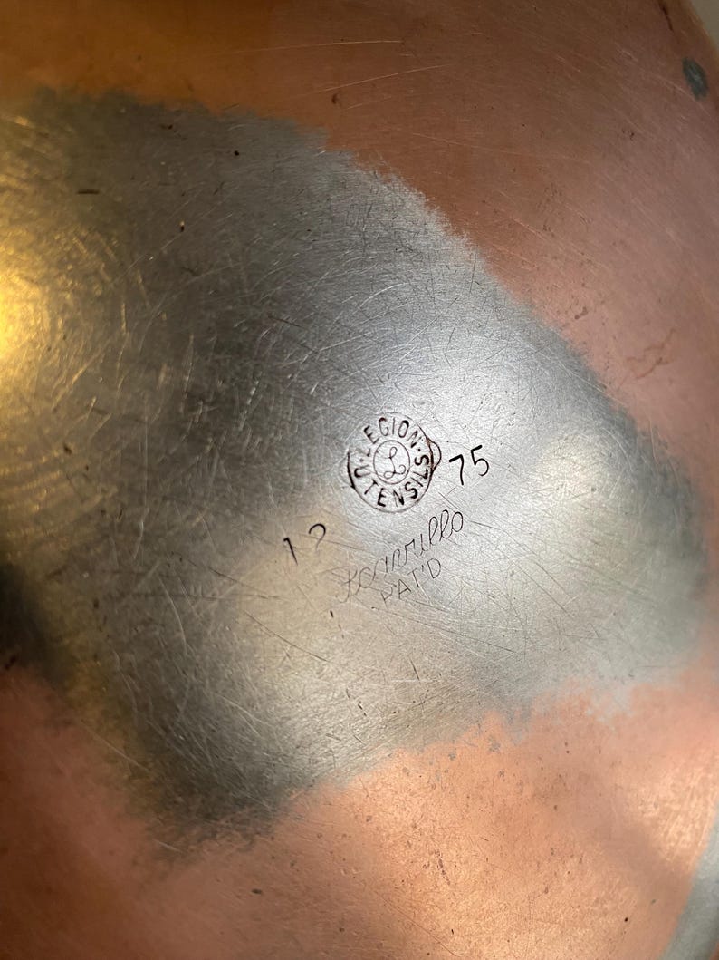 May include: A copper-coloured metal bowl with a brushed finish. The bowl features a circular stamp with the text "LEGIONS UTENSILS" and the number "75". Additional text includes "12" and "Scorrillo PAT'D".