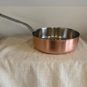 May include: A copper saucepan with a stainless steel interior and a long, silver handle. The pan is on a beige fabric surface. The cookware is designed for stovetop use.