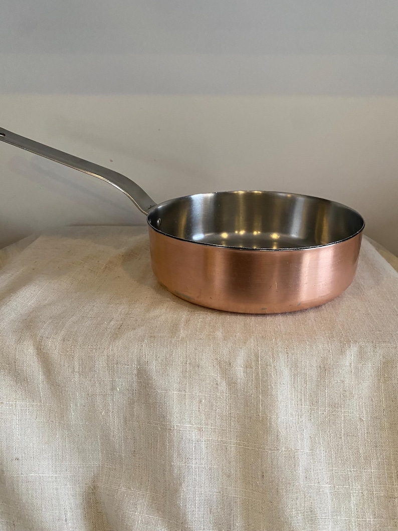 May include: A copper saucepan with a stainless steel interior and a long, silver handle. The pan is on a beige fabric surface. The cookware is designed for stovetop use.