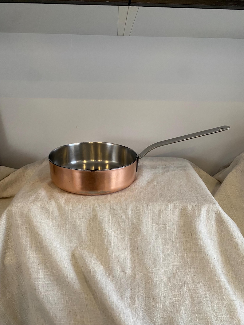 May include: A copper-bottomed saucepan with a stainless steel interior and a long, silver handle. The round pan sits on a light-colored, textured surface. The handle extends to the right.