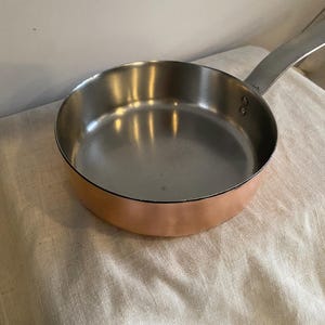 May include: A copper-bottomed frying pan with a stainless steel interior and a long, riveted handle. The pan reflects light, showing its polished surface. The pan is designed for cooking.