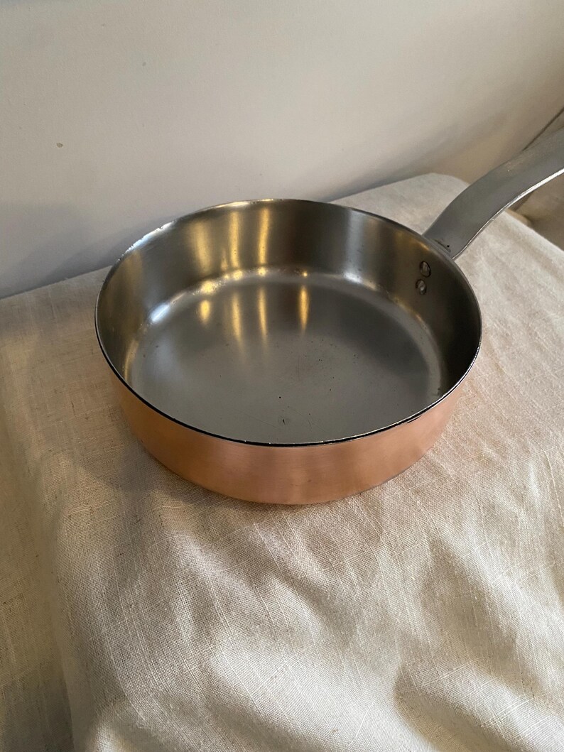 May include: A copper-bottomed frying pan with a stainless steel interior and a long, riveted handle. The pan reflects light, showing its polished surface. The pan is designed for cooking.