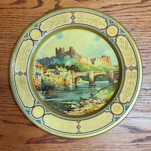 May include: A round metal decorative plate featuring a colorful landscape illustration of Richmond Castle with a stone bridge over a river. The border is decorated with gold and purple filigree patterns on a light yellow background.