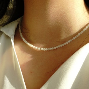 Peut inclure: Un collier de perles blanches avec un design délicat. Le collier est composé de petites perles rondes et est porté par une personne aux cheveux longs et foncés.