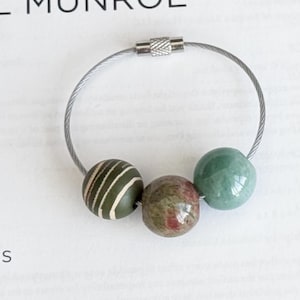 Puede incluir: Un llavero de metal plateado con tres cuentas decorativas. Las cuentas son una mezcla de colores y patrones: una verde oliva con rayas blancas, una moteada roja y marrón y una verde lisa.
