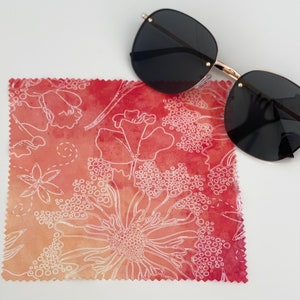 Könnte beinhalten: Ein rotes und oranges Mikrofaser-Reinigungstuch mit einem weißen Blumenmuster. Das Tuch ist in der Mitte gefaltet und hat einen gewellten Rand. Eine schwarze Sonnenbrille mit goldenen Bügeln liegt auf dem Tuch.