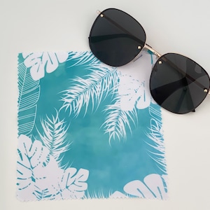 Puede incluir: Un paño de microfibra azul turquesa con un patrón de hojas tropicales blancas. El paño está doblado por la mitad y un par de gafas de sol negras están encima.