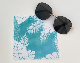 Puede incluir: Un paño de microfibra azul turquesa con un patrón de hojas tropicales blancas. El paño está doblado por la mitad y un par de gafas de sol negras están encima.