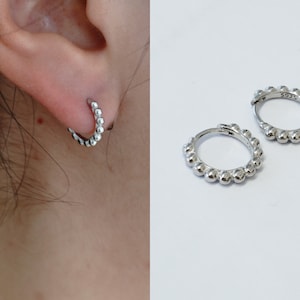 May include: A pair of small, silver hoop earrings. Each earring features a series of small, round beads along the outer edge. One earring is shown on an ear, while the other is displayed separately. The earrings have a shiny, metallic finish. The earrings are marked with 'S925'.